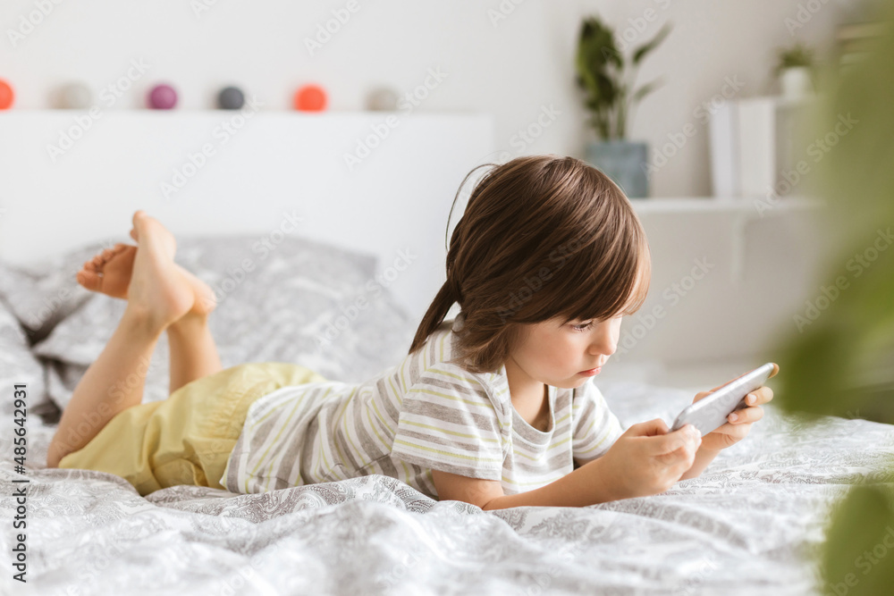 A little boy of four years lies on the bed and plays a mobile phone, a smartphone. Children use technology. The problem of children and gadgets A little boy of four years lies on the bed and plays a mobile phone, a smartphone. Children use technology. The problem of children and gadgets