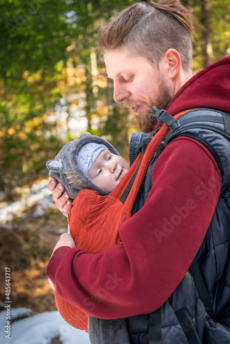 Side view of young bearded babywearing father with his baby in baby carrier walking outdoor