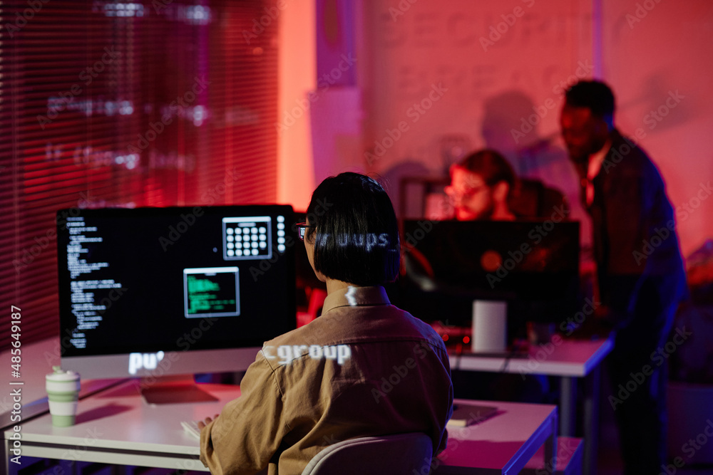Rear view of young contemporary programmer sitting in front of computer screen with decoded data and working in openspace office Rear view of young contemporary programmer sitting in front of computer screen with decoded data and working in openspace office