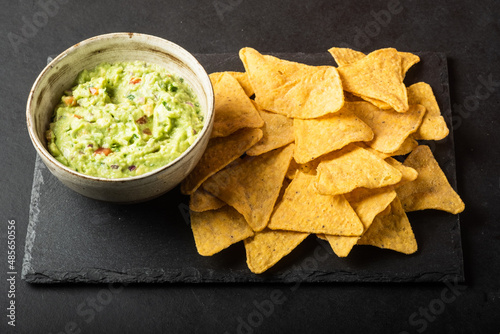 Mexican nachos chips and bowl with homemade fresh guacomole sauce made from avocado and lime  over black background.
