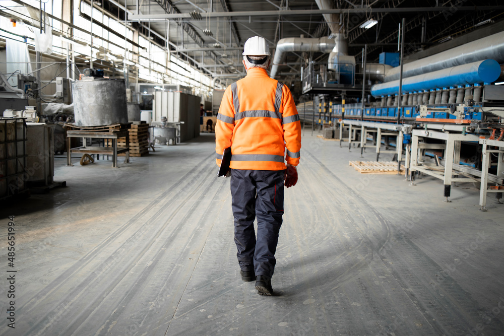 Professional factory worker or engineer in high visibility jacket and hardhat walking through industrial production hall with machinery. Factory interior.