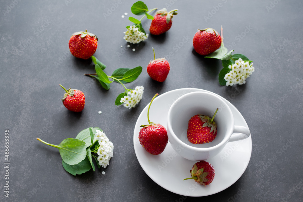 White spirea flowers and bright red strawberries in a white cup and scattered around on a black background. Summer mood background