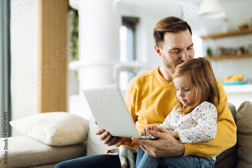 Young father and his small daughter using computer on sofa at home