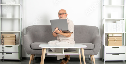 Mature consultant, senior man in office. Old man sits at desk on sofa with laptop, portrait. Older senior business man, grey-haired man wearing glasses looking at laptop. © Volodymyr