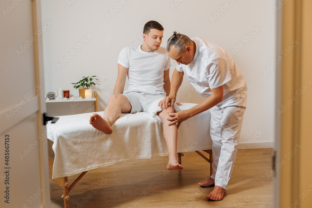 Handsome masseur therapist in white uniform making manual therapy for young athlete knee. Professional massage treatment and rehabilitation for sportsmen. Concept of wellness, body and health care. Handsome masseur therapist in white uniform making manual therapy for young athlete knee. Professional massage treatment and rehabilitation for sportsmen. Concept of wellness, body and health care.