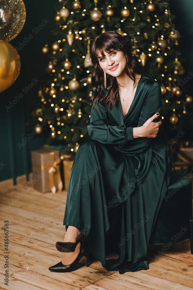 A beautiful pregnant brunette girl in a green dress sits against the background of balloons and a Christmas tree.
