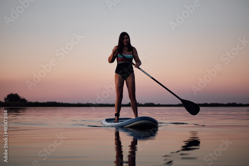 Female sup boarder holding oar in hands rowing looking at camera on calm lake with stunning pink sky in background in swimsuit in summer. Active lifestyle for older people.