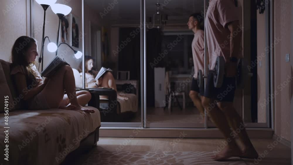 Lunges with dumbbells. A man trains his legs in an apartment in front of a girl with a book Lunges with dumbbells. A man trains his legs in an apartment in front of a girl with a book