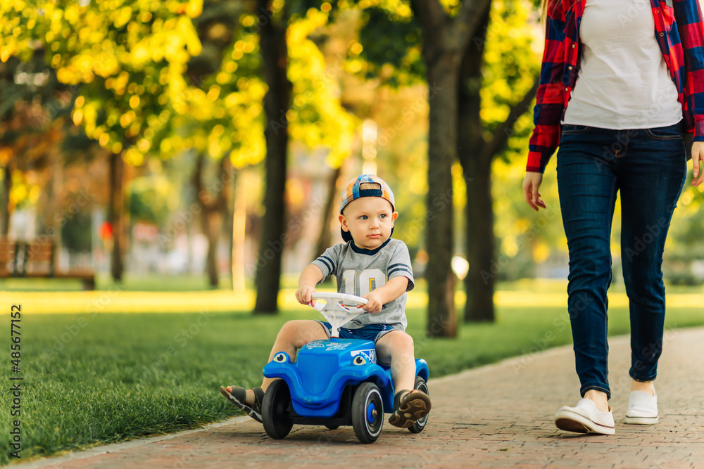 Mom teaches her son to ride a toy car in the park. Preschooler driving his toy car outdoors in summer Mom teaches her son to ride a toy car in the park. Preschooler driving his toy car outdoors in summer