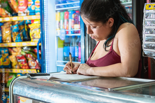 latina woman concentrating on writing in her notebook the accounting of the neighborhood store, girl leaning on a glass display case thinking about sales.