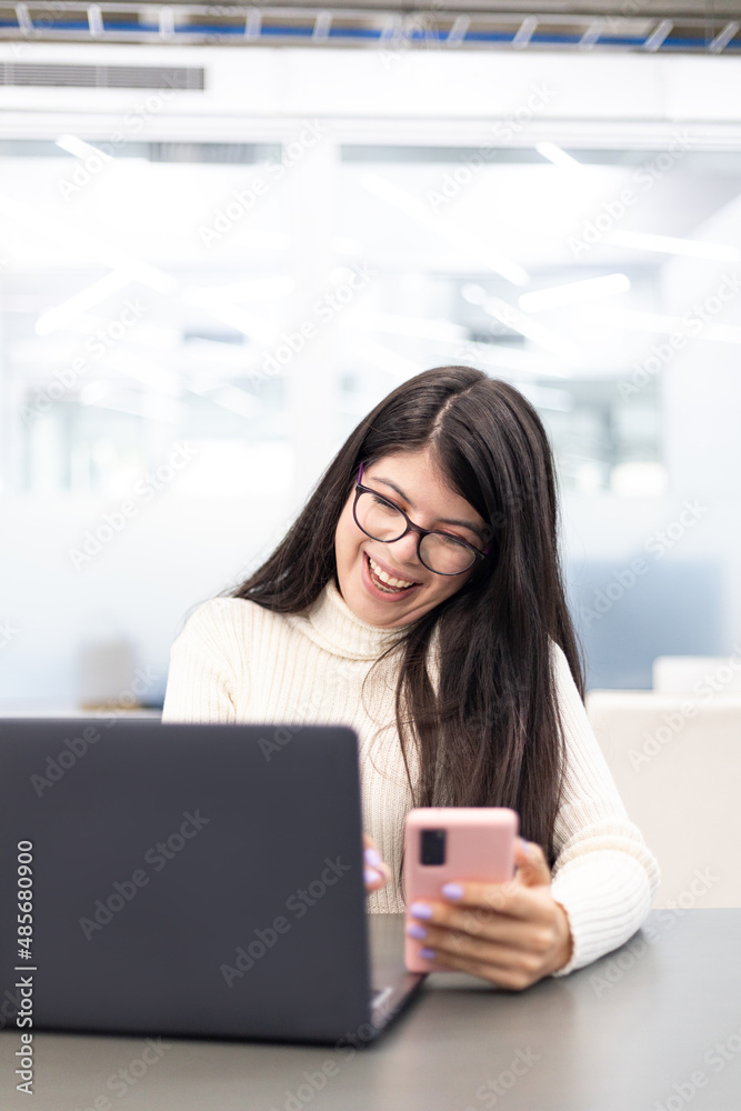 White young woman with happy expression using a laptop and cellphone on a desk looking to the screen White young woman with happy expression using a laptop and cellphone on a desk looking to the screen
