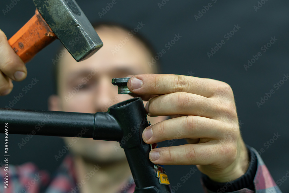 A bicycle mechanic with a hammer in his hands on a black background. The mechanic is holding an old frame in his hands, replacing the headset and cartridge cup bearings. Steering tube.