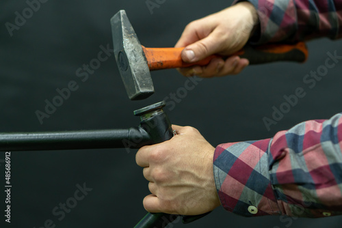 A bicycle mechanic with a hammer in his hands on a black background. The mechanic is holding an old frame in his hands, replacing the headset and cartridge cup bearings. Steering tube.