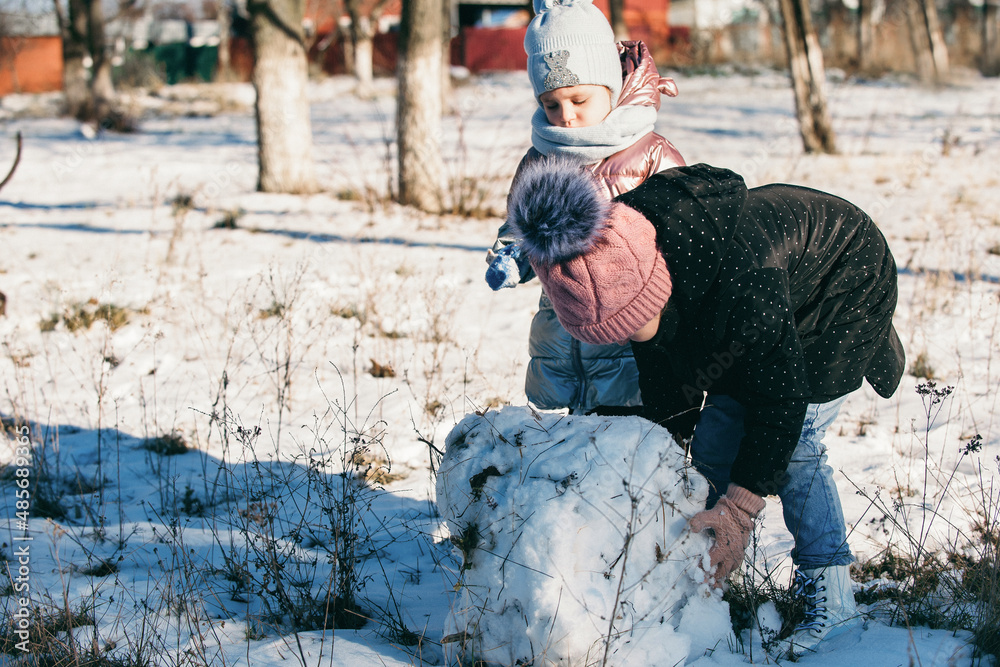 Two sisters actively and have fun on a winter walk, they roll a snowball and build a snowman Two sisters actively and have fun on a winter walk, they roll a snowball and build a snowman