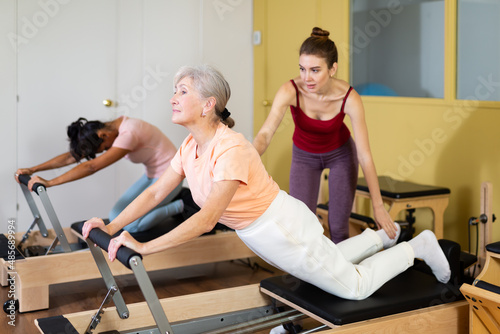 Focused positive elderly woman practicing pilates on reformer to improve and maintain mobility under supervision of qualified young female trainer photo