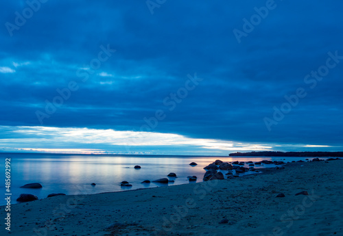 Long exposure night scenery of the Onega lake shore. Sand and stones on the beach, dark blue clouds in the sky, reflection of the last orange sunset rays in the water