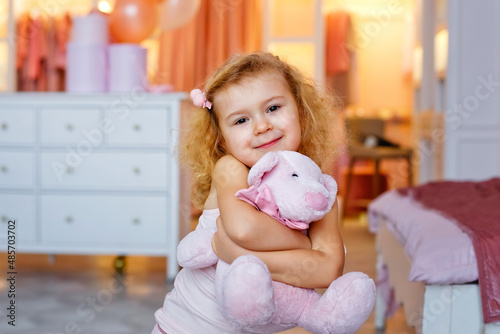 Portrait of a small curly blonde with a stuffed toy in the bedroom. A girl hugs a pink bear.