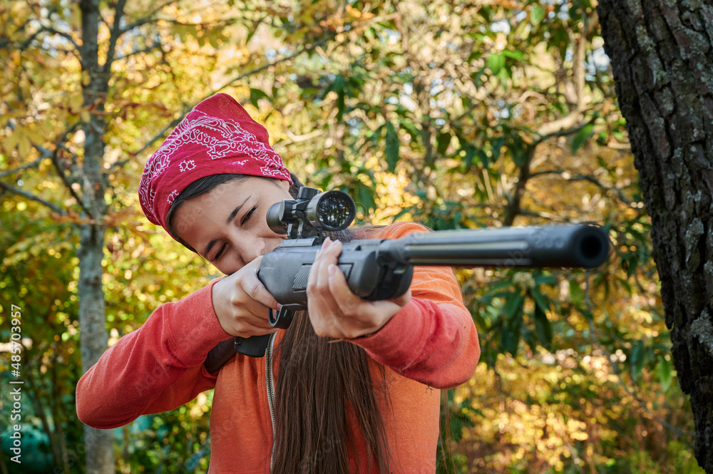 Sideview of teenage woman dressed in colorful clothing wields a shotgun while aiming using a telescopic sight. Learning to hunt in the forest.