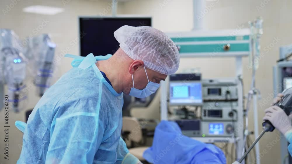 Doctor bending low over the patient in the process of operation. Assistant’s hands holding instrument for surgery. Operation room in blur at backdrop. Doctor bending low over the patient in the process of operation. Assistant’s hands holding instrument for surgery. Operation room in blur at backdrop.
