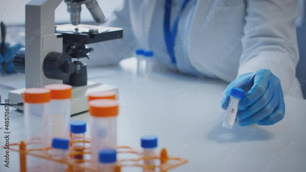 Close up of scientist wearing white coat looking through microscope and making notes whilst holding test tube which he puts on desk to show label pi - shot in slow motion Close up of scientist wearing white coat looking through microscope and making notes whilst holding test tube which he puts on desk to show label pi - shot in slow motion