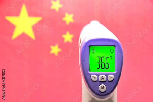 Concept of winter Games in 2022 in Beijing, China, due to threat of new coronavirus infection. Infrared thermometer shows normal healthy body temperature on blurred background of red Chinese flag