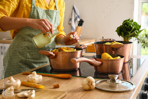 Woman in apron adding oil from bottle to cooking pot with vegetable stew in kitchen