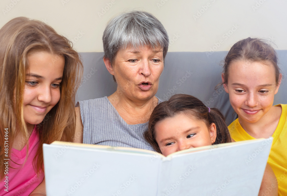 Grandmother reads a book to her granddaughters sitting together Grandmother reads a book to her granddaughters sitting together