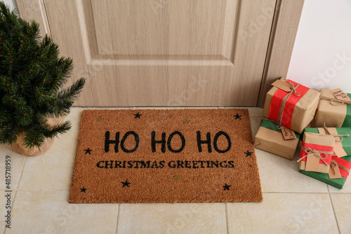 Mat with Christmas greeting, gifts and small fir tree near light wooden door