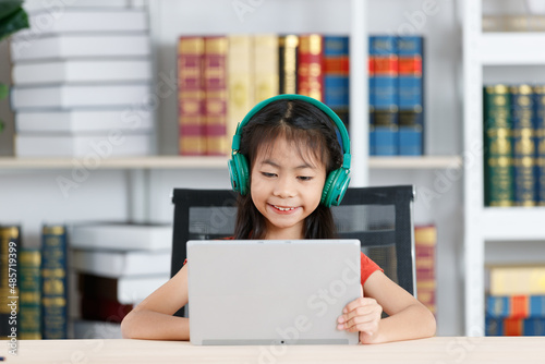 Asian clever smart diligent young primary school girl daughter in casual outfit wearing big headphone smiling using touchscreen tablet computer in hands learning studying online during quarantine