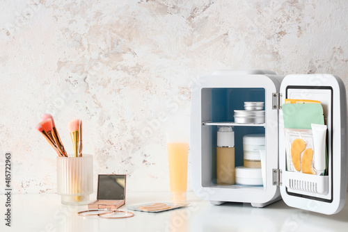 Quadro em tela Small cosmetic refrigerator with products and makeup brushes on table near light