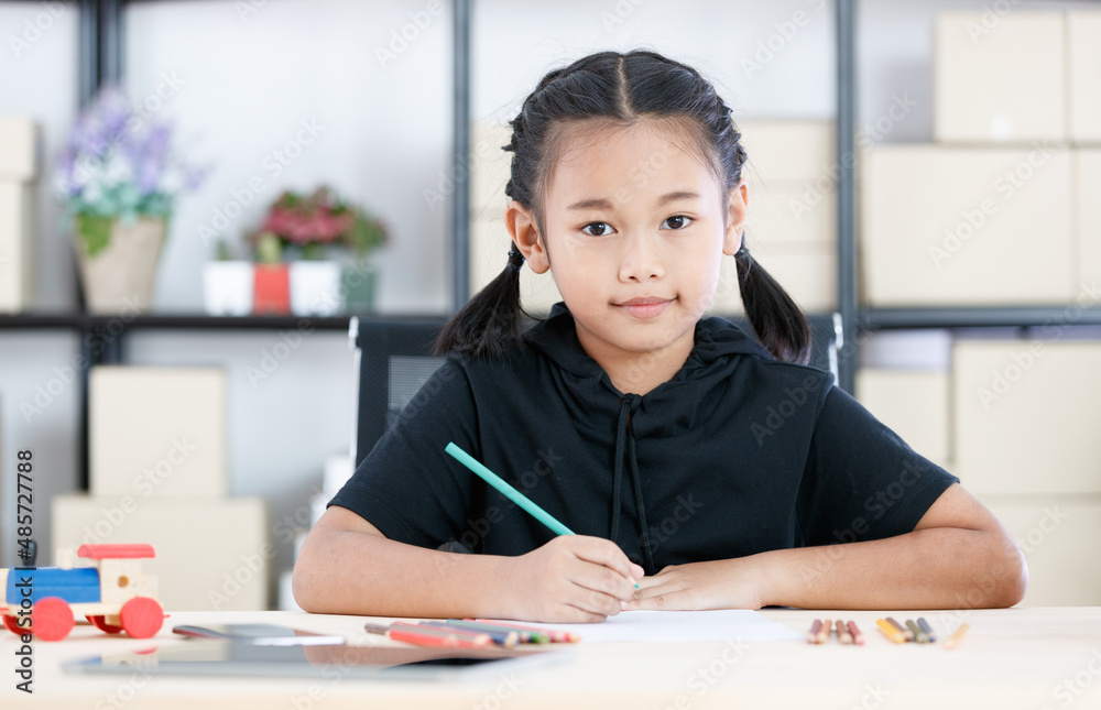 Asian young smart clever elementary schoolgirl artist in casual outfit sitting using color pencils drawing painting picture on paper alone on working table with tablet and wooden toy in living room Asian young smart clever elementary schoolgirl artist in casual outfit sitting using color pencils drawing painting picture on paper alone on working table with tablet and wooden toy in living room