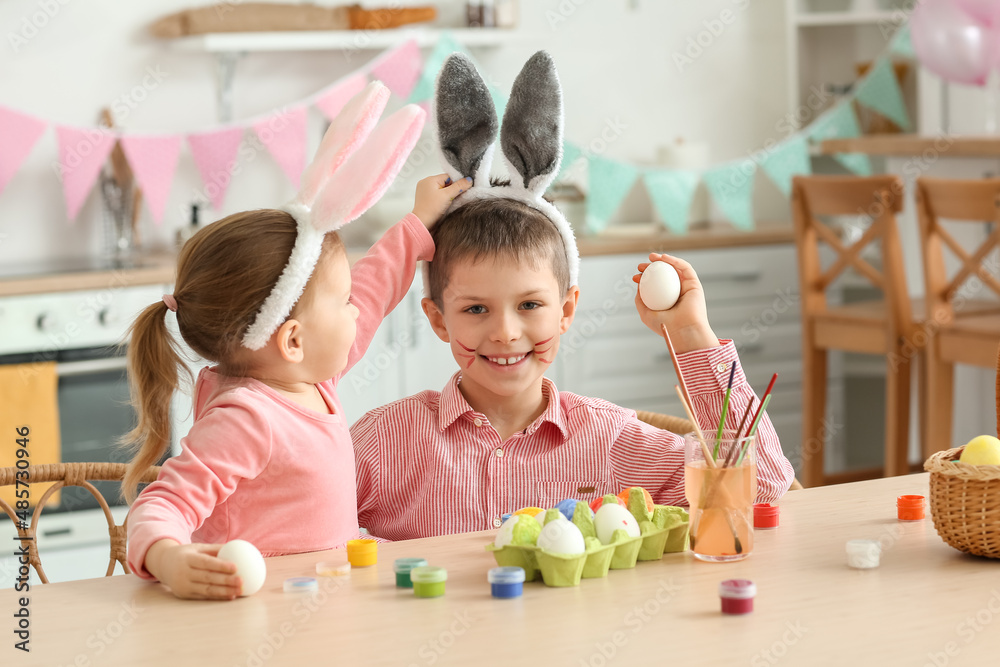 Fototapeta premium Cute little girl in bunny ears with her brother painting Easter eggs at home