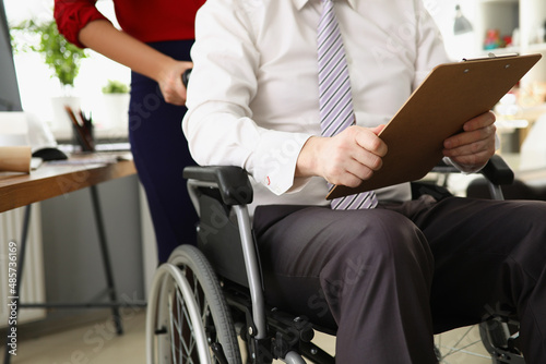 Man sit in wheelchair with clipboard, woman drive boss in office