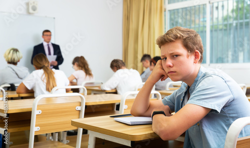 Portrait of disgruntled tired teenager male student sitting at table in classroom during lesson