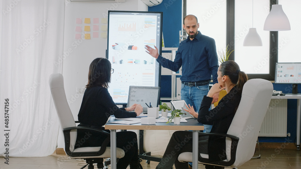 Man pointing at television with chart presentation for project planning while businesswomen listening to ideas for marketing strategy at meeting. Workmates with research development Man pointing at television with chart presentation for project planning while businesswomen listening to ideas for marketing strategy at meeting. Workmates with research development