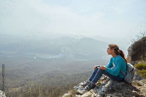 A girl in the mountains sits on a rock and looks at the surrounding landscape. Active sports and recreation.