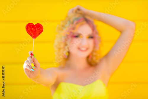 Beautiful young happy girl with colorful summer rainbow makeup tries heart shaped lollipop. Emotions of happiness and fun. Summer holidays, playful mood at a party. Emotions of love.