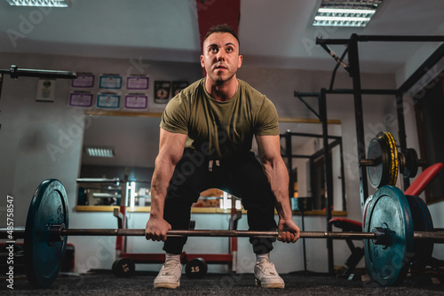 One man young adult caucasian male bodybuilder training back doing deadlift with barbel and weights while standing in the gym wearing shirt dark photo real people copy space front view full length