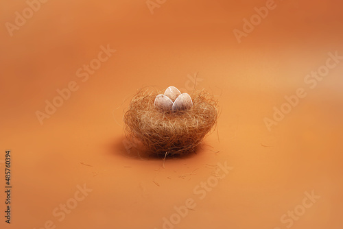 A small bird's nest made of dry grass with three painted white eggs inside on an orange, brown, yellow isolated background, side view 