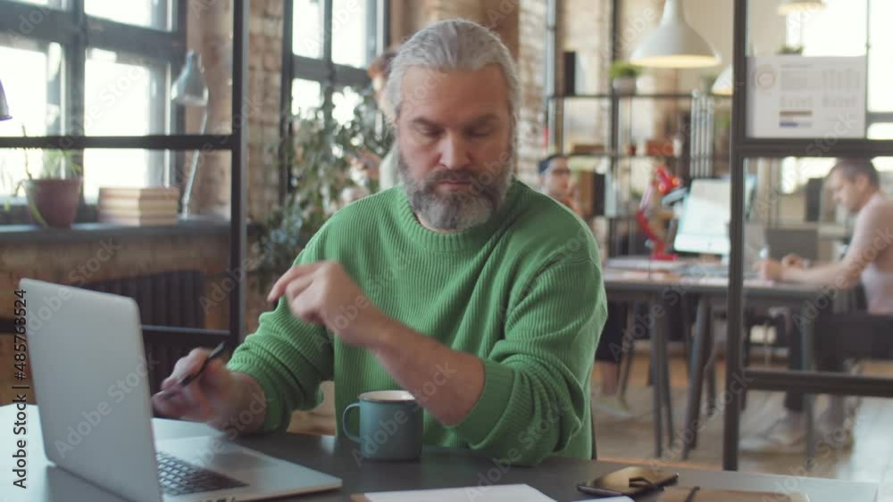 Mature businessman typing on laptop and checking documents while working at desk in modern open office Mature businessman typing on laptop and checking documents while working at desk in modern open office