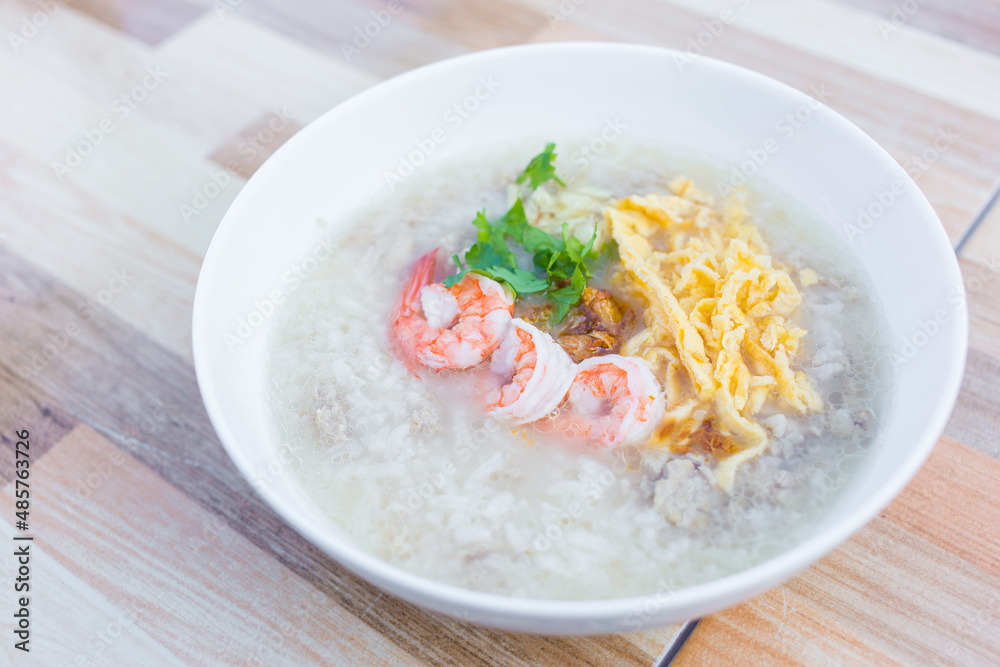 Boiled rice with pork was added on top with chopped omelet, boiled shrimp and coriander as a a breakfast meal in the morning in Thai style while that bowl put on wooden background. Boiled rice with pork was added on top with chopped omelet, boiled shrimp and coriander as a a breakfast meal in the morning in Thai style while that bowl put on wooden background.