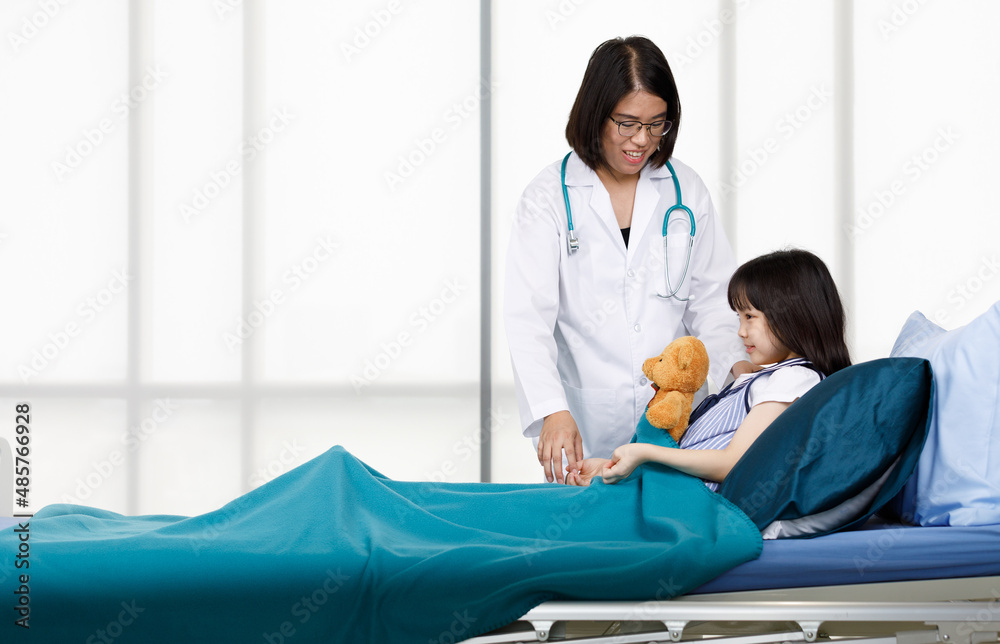 Asian professional friendly female doctor in white lab coat and stethoscope standing smiling to young girl patient lay down on bed hugging teddy bear doll in hospital wardroom Asian professional friendly female doctor in white lab coat and stethoscope standing smiling to young girl patient lay down on bed hugging teddy bear doll in hospital wardroom