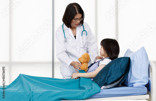 Asian professional friendly female doctor in white lab coat and stethoscope standing smiling to young girl patient lay down on bed hugging teddy bear doll in hospital wardroom