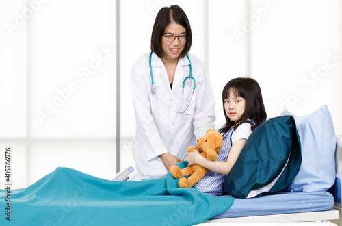 Asian professional friendly female doctor in white lab coat and stethoscope standing smiling to young girl patient lay down on bed hugging teddy bear doll in hospital wardroom photo