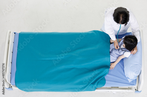 Top view portrait shot of Asian professional friendly female doctor in white lab coat and stethoscope standing smiling holding comforting young recovery girl patient laying down on bed in hospital