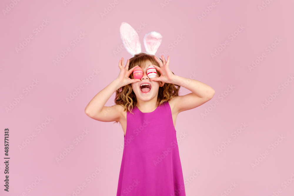 Adorable cheerful little girl with curly hair in fuchsia colored dress and bunny ear headband, smiling happily while covering eyes with eggs during Easter celebration