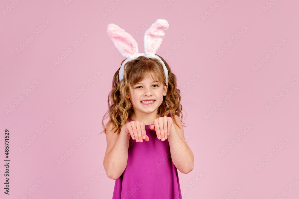 Adorable funny little kid with curly blond hair in dress and bunny ears, smiling and showing paws gesture while imitating rabbit during Easter party in pink studio Adorable funny little kid with curly blond hair in dress and bunny ears, smiling and showing paws gesture while imitating rabbit during Easter party in pink studio