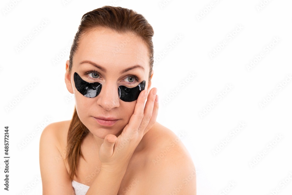 a woman applies cosmetic black patches under her eyes against a white wall a woman applies cosmetic black patches under her eyes against a white wall