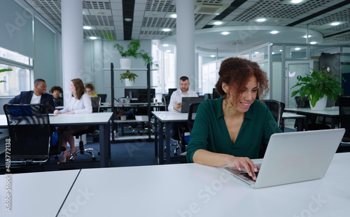 Black businesswoman using laptop in open space office, smiling during online communication, diverse coworkers on blurred background. Company employee in shared working environment