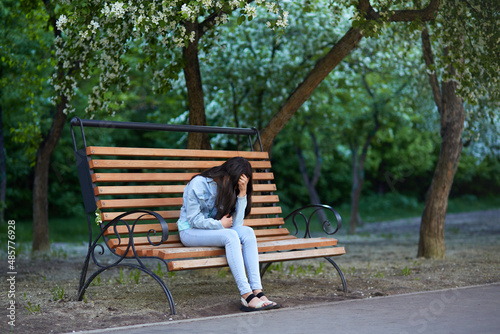 Upset sad teenage girl sitting on a bench in the park, holding her head, face covered. Mental health. © Viktoriya